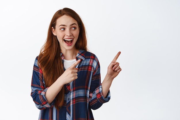 Cheerful young woman with red long hair, smiling and laughing, show fantastic news, pointing aside right and looking at advertisement, click on link, white background