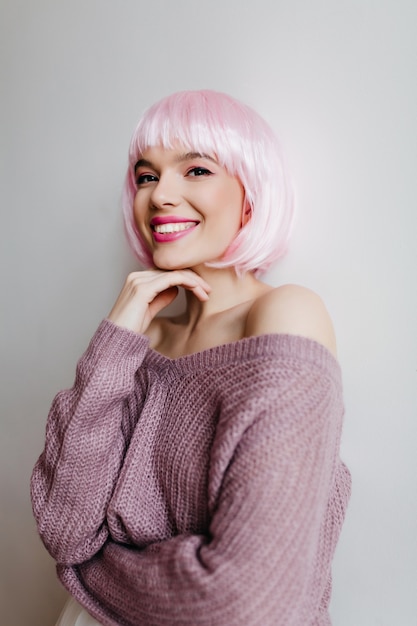 Веселая молодая женщина с блестящими светло-розовыми волосами, улыбаясь на светлой стене. Крытый портрет великолепной женской модели в милом перуке и фиолетовом свитере.