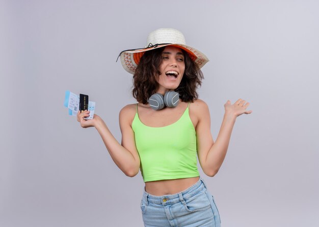 Веселая молодая женщина с короткими волосами в зеленом топе в наушниках в шляпе от солнца держит билеты на самолет и кредитную карту на белом фоне