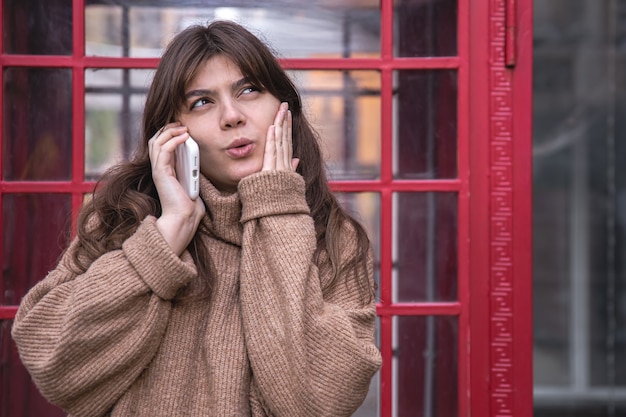 Free photo cheerful young woman with a smartphone on the background of a telephone booth