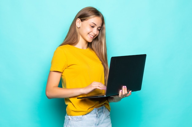 Веселая молодая женщина, работающая на ноутбуке, позирует изолированной на бирюзовой стене
