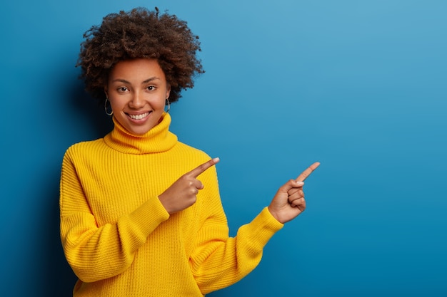 cheerful young woman in yellow sweater points aside at copy space, shows cool advertisement against blue background