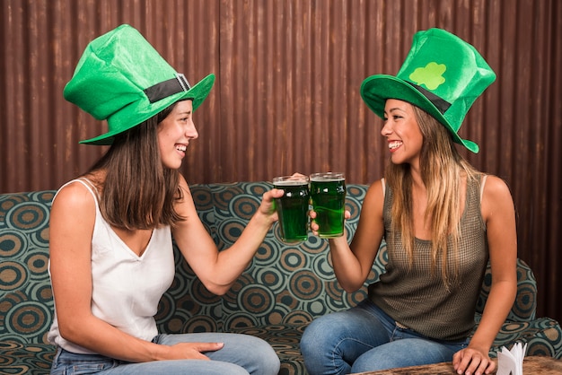 Cheerful young women clanging glasses of drink on settee in room