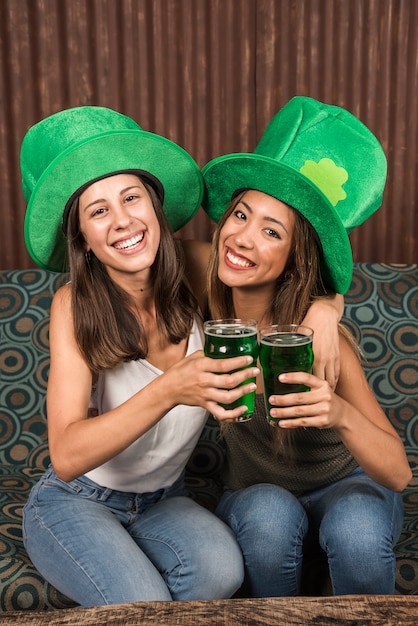 Веселые молодые женщины, обнимающие и звенящие бокалами с напитком на диване