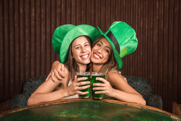 Cheerful young women hugging with glasses of drink on settee near table