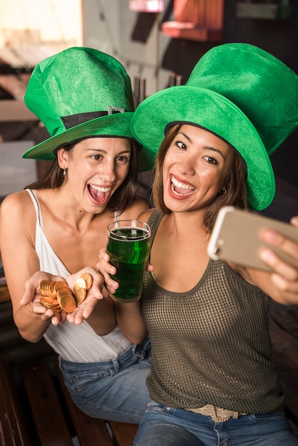 Cheerful young women with glass of drink and golden coins taking selfie on smartphone