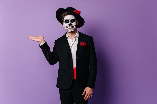 Cheerful zombie boy standing in studio with smile. Indoor photo of funny european man with muerte makeup isolated on purple background.