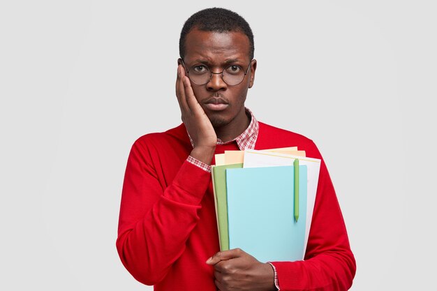 Cheerless black man has dejected facial expression, keeps hand on cheek, feels tired of studying, holds textbooks with pen, going to library