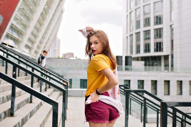 Cheery girl in bright attire looking over shoulder with interest walking on urban street