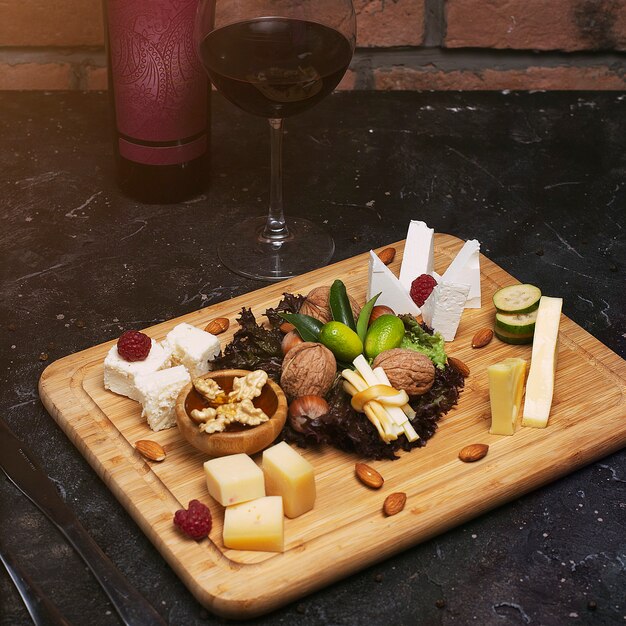 さまざまなチーズ、ぶどう、ナッツ、蜂蜜、パン、素朴な木のナツメヤシとチーズの盛り合わせ。ワインのボトルとワインのグラスと暗い木の板に