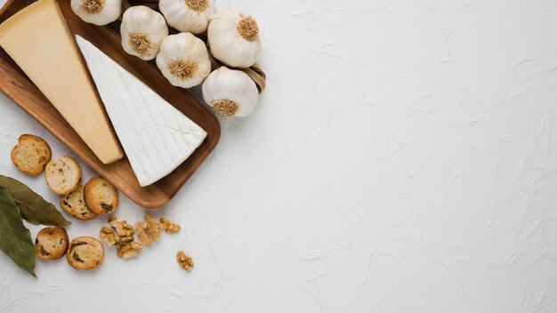 Cheeses piece on wooden tray with bay leaves; bread slice; walnut and garlic bulb on white surface