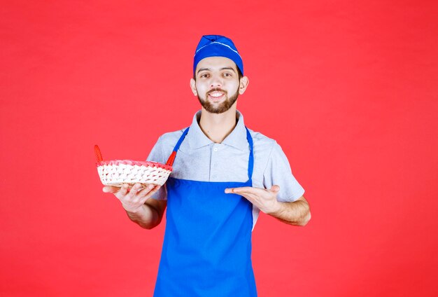 Шеф-повар в синем фартуке держит корзину с хлебом, накрытую красным полотенцем.