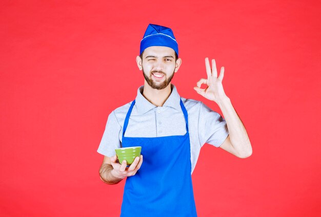 Chef in blue apron holding a green ceramic cup and showing satisfaction sign. 