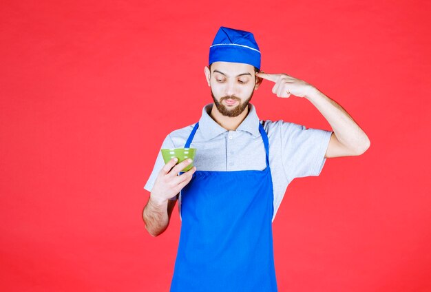 Chef in blue apron holding a green ceramic cup and thinking over it. 