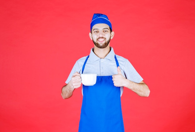 Free photo chef in blue apron holding a white ceramic cup and showing enjoyment sign.