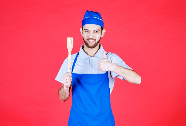 Chef in blue apron holding a wooden spatula and showing enjoyment sign. 