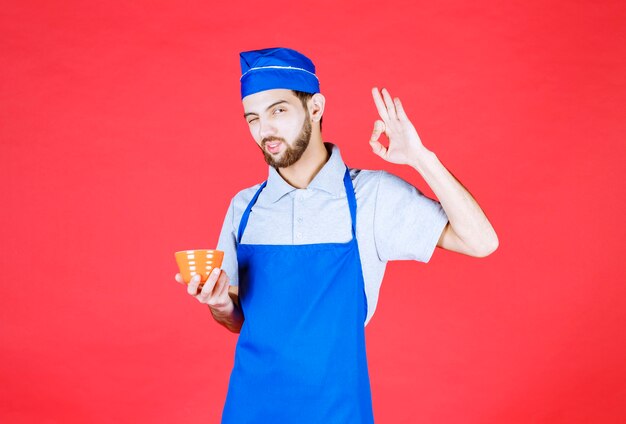 Chef in blue apron holding a yellow ceramic cup and showign enjoyment sign. 