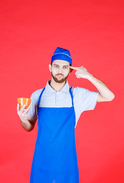 Chef in grembiule blu che tiene una tazza di ceramica gialla e pensa.