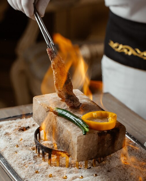 шеф-повар готовит кусок говяжьего стейка с зажимом на каменном кирпиче в огне