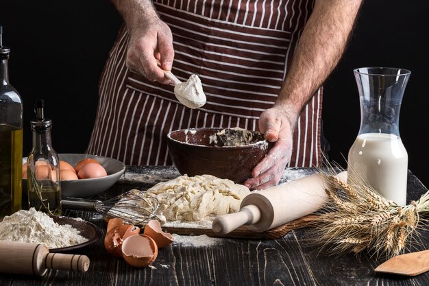 Chef hands are sprinkling flour into bowl to make dough. Close up view on black background