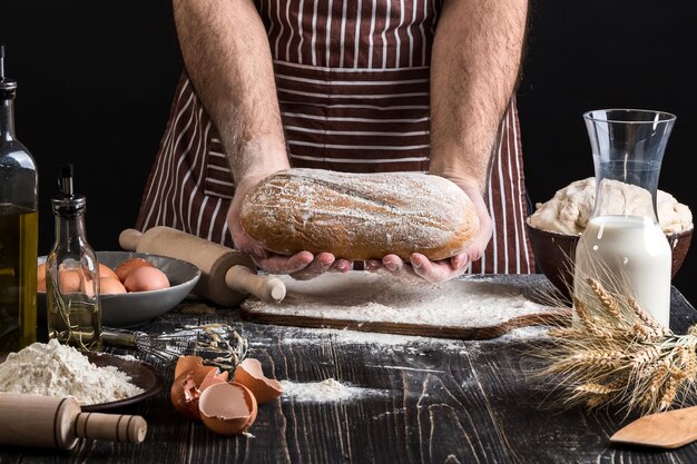 Chef holds the fresh bread in hand. Man preparing dough at table in kitchen. On black background. Healthy or cooking concept.
