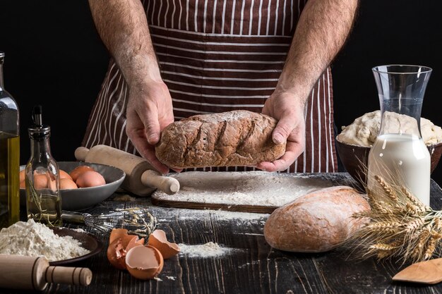 Chef holds the fresh bread in hand. Man preparing dough at table in kitchen. On black background. Healthy or cooking concept.