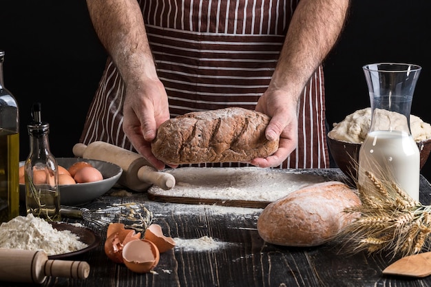 Шеф-повар держит в руке свежий хлеб. Мужчина готовит тесто за столом на кухне. На черном фоне. Здоровая или кулинарная концепция.