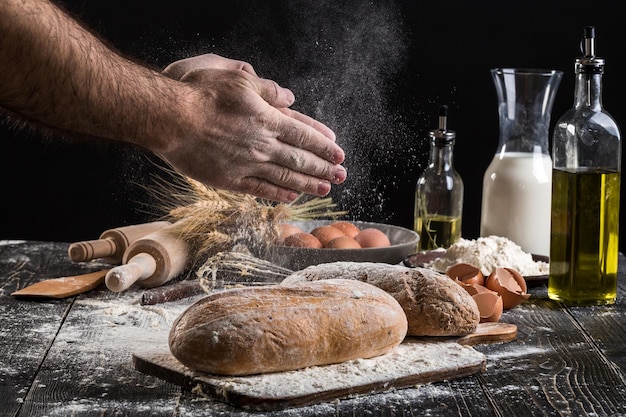 Chef sprinkles fresh bread with flour. Man preparing dough at table in kitchen. On black background. Healthy or cooking concept.