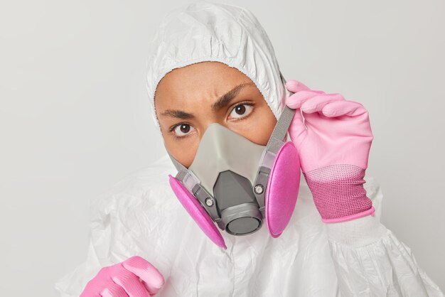 Химическая или биологическая безопасность Внутренний снимок серьезной женщины в радиационно-защитном костюме с капюшоном и респиратором серьезно смотрит на камеру, изолированную на белом фоне Профессиональное дезинфицирующее средство