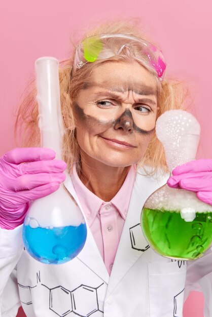  chemist conducts scientific research holds two glass flasks with blue and green liquid makes experiment in laboratory wears uniform isolated on pink 