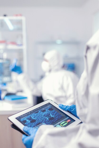 Chemist dressed in protection suit for coronavirus holding tablet pc in laboratory. Team of scientists conducting vaccine development using high tech technology for researching treatment against covid