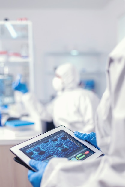 실험실에서 태블릿 PC를 들고 있는 코로나바이러스 보호복을 입은 화학자. covid에 대한 치료법을 연구하기 위해 첨단 기술을 사용하여 백신 개발을 수행하는 과학자 팀