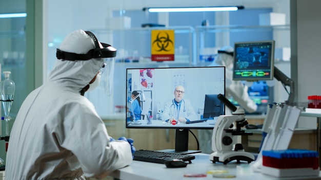 PPE 정장을 입은 화학자는 화상 통화로 전문 의사를 듣고 연구 실험실에서 가상 회의 중에 토론합니다. covid19 바이러스에 대한 치료법을 연구하기 위해 첨단 기술을 사용하는 의사