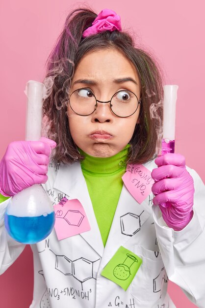 химик в белом медицинском халате впервые проводит эксперимент держит пробирку и стеклянную колбу с цветной жидкостью работает в научной лаборатории дует щеки позы на розовый