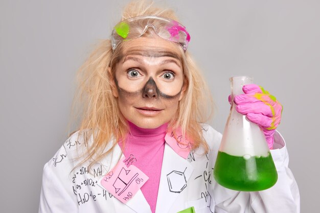 化学実験の予想外の結果に呆然とした目の周りの汚れを持った化学者は、白衣を着た緑色の液体が入ったガラス製フラスコを屋内に持っています。生化学スペシャリスト