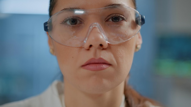 무료 사진 화학자 여성은 안전과 보호를 위해 안경을 쓰고 과학 실험실에서 카메라를 보고 있습니다. 실험실에서 작업하는 보호용 고글을 쓴 생물학 연구원의 초상화. 확대