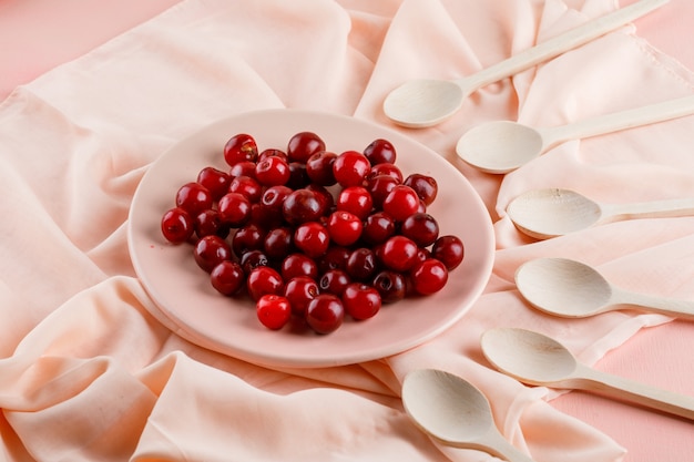 Бесплатное фото Вишня в тарелке с деревянными ложками высокого угла зрения на текстиле и розовый