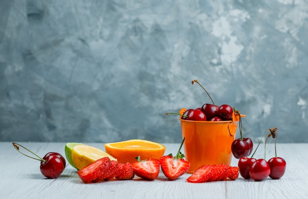Cherries in a mini bucket with lemon, lime, orange, strawberries side view on wooden and grunge background