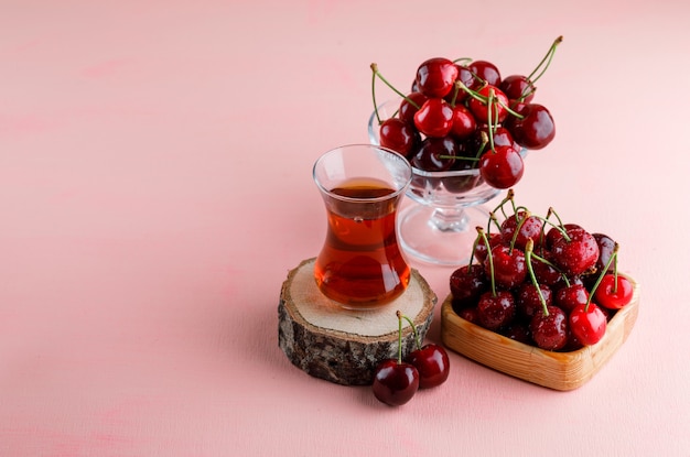 Cherries with glass of tea on wooden board in wooden plate and vase on pink surface