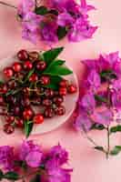 Бесплатное фото Вишня с листьями, цветочными ветками в тарелке на розовой поверхности
