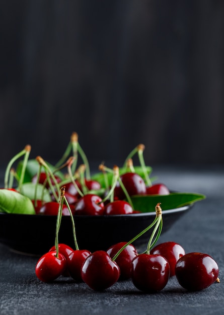 Cherries with leaves in a plate on grey and dark surface, side view