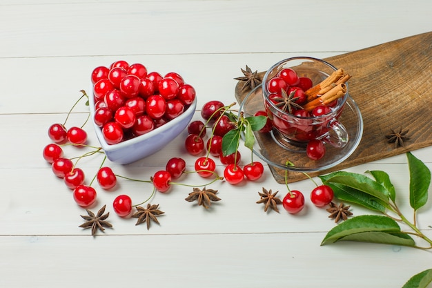 Free photo cherries with leaves, spices in bowl and glass on wooden and cutting board, high angle view.