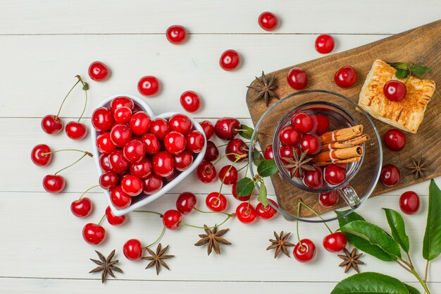 Cherries with pastry, leaves, spices in bowl and glass on wooden and cutting board.