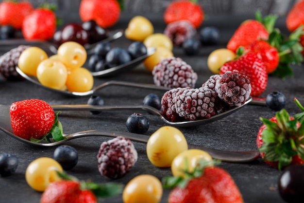 Cherries with strawberries, blueberries, mulberries in spoons on dark table, high angle view.