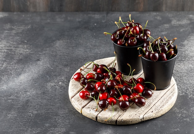 Cherries with wooden board in buckets on grungy and wooden surface, high angle view.