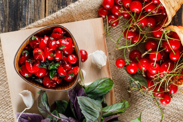 Cherry in baskets with garlic, jam, basil, cutting board on wooden and piece of sack