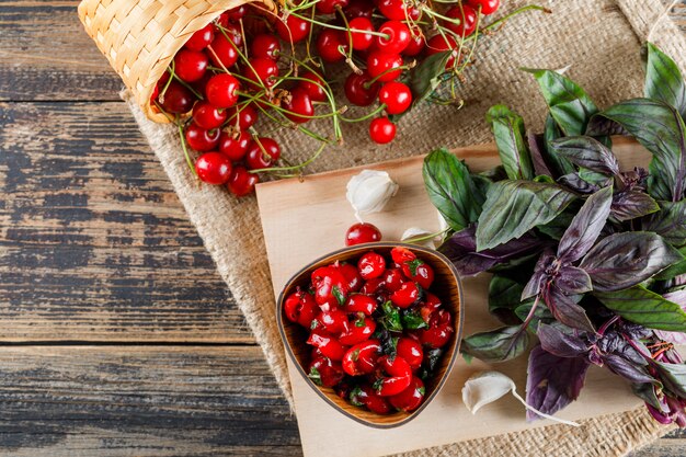 Cherry in baskets with jam, garlic, basil, cutting board top view on wooden and piece of sack