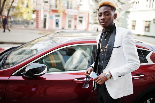 Elegante uomo afroamericano bello in abito bianco contro un'auto di lusso rossa con il telefono cellulare a portata di mano