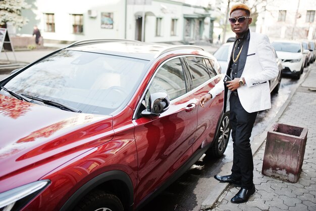 手元に携帯電話と赤い高級車に対して白いスーツでシックなハンサムなアフリカ系アメリカ人の男