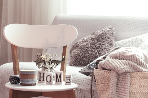 家のシックなインテリア。キャンドル、白い木製の椅子に家の木製の文字が付いている花が付いているつぼ。バックグラウンドにクッションが付いたソファーと籐のバスケット。家の装飾。